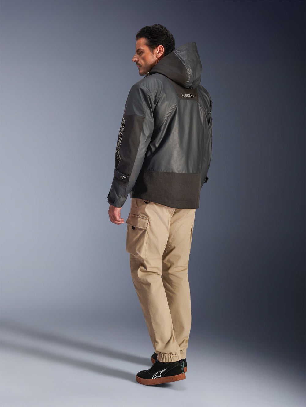 Man wearing the Alpinestars Mospher Waterproof Jacket in Black, back and side profile view, paired with tan cargo pants and black sneakers. Features a hooded parka design with ripstop fabric, Alpinestars branding on the sleeve, and a modern urban motorcycle style.
