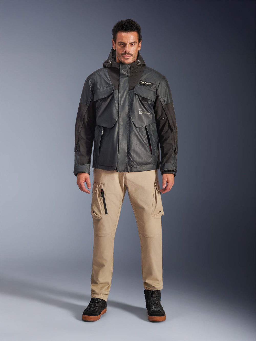 Male model wearing the Alpinestars Mospher Waterproof Jacket in Black, urban contemporary motorcycle gear, tactical utility design with multiple functional pockets and hood, paired with tan cargo pants and black shoes, full-body studio portrait.