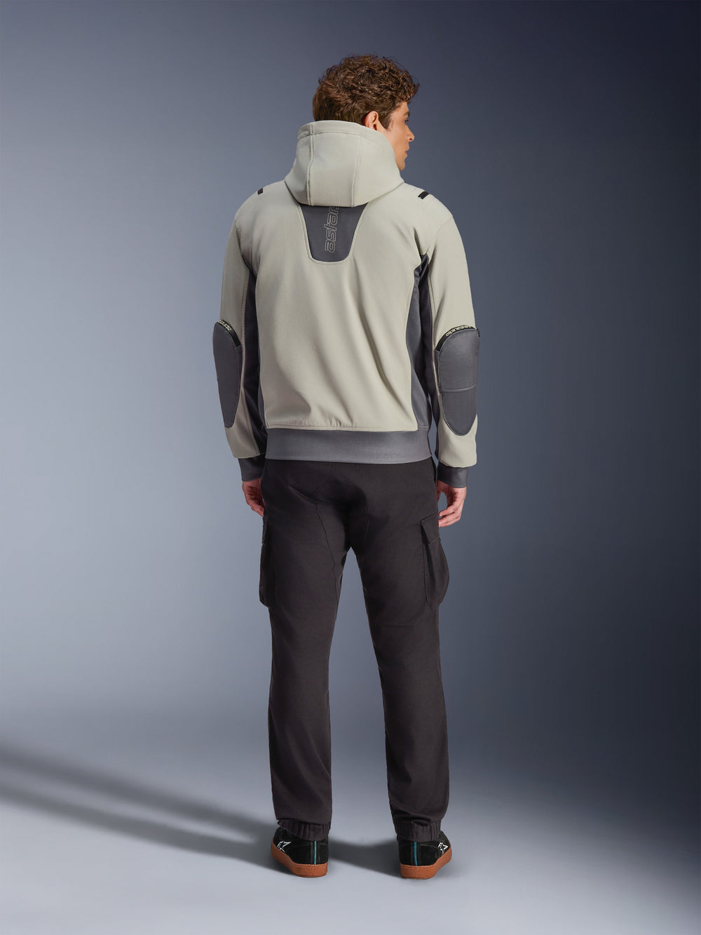 Alpinestars Moblast Waterproof Jacket, Iron Gray Dark Sage, rear view of a male rider wearing the hooded motorcycle jacket, light gray and dark sage colorway, vertical 'astars' logo on the upper back, dark gray elbow patches, styled with dark cargo pants for an urban contemporary look.