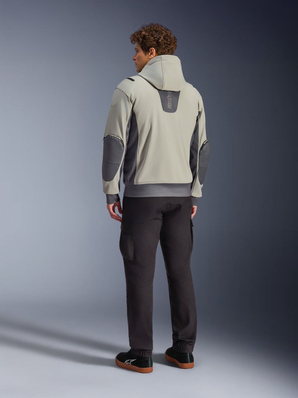 Male model viewed from the back wearing the Alpinestars Moblast Waterproof Jacket in Iron Gray Dark Sage, hooded urban motorcycle jacket with dark gray side panels and reinforced elbow patches, paired with dark riding pants and black sneakers, modern minimalist aesthetic for urban commuting.