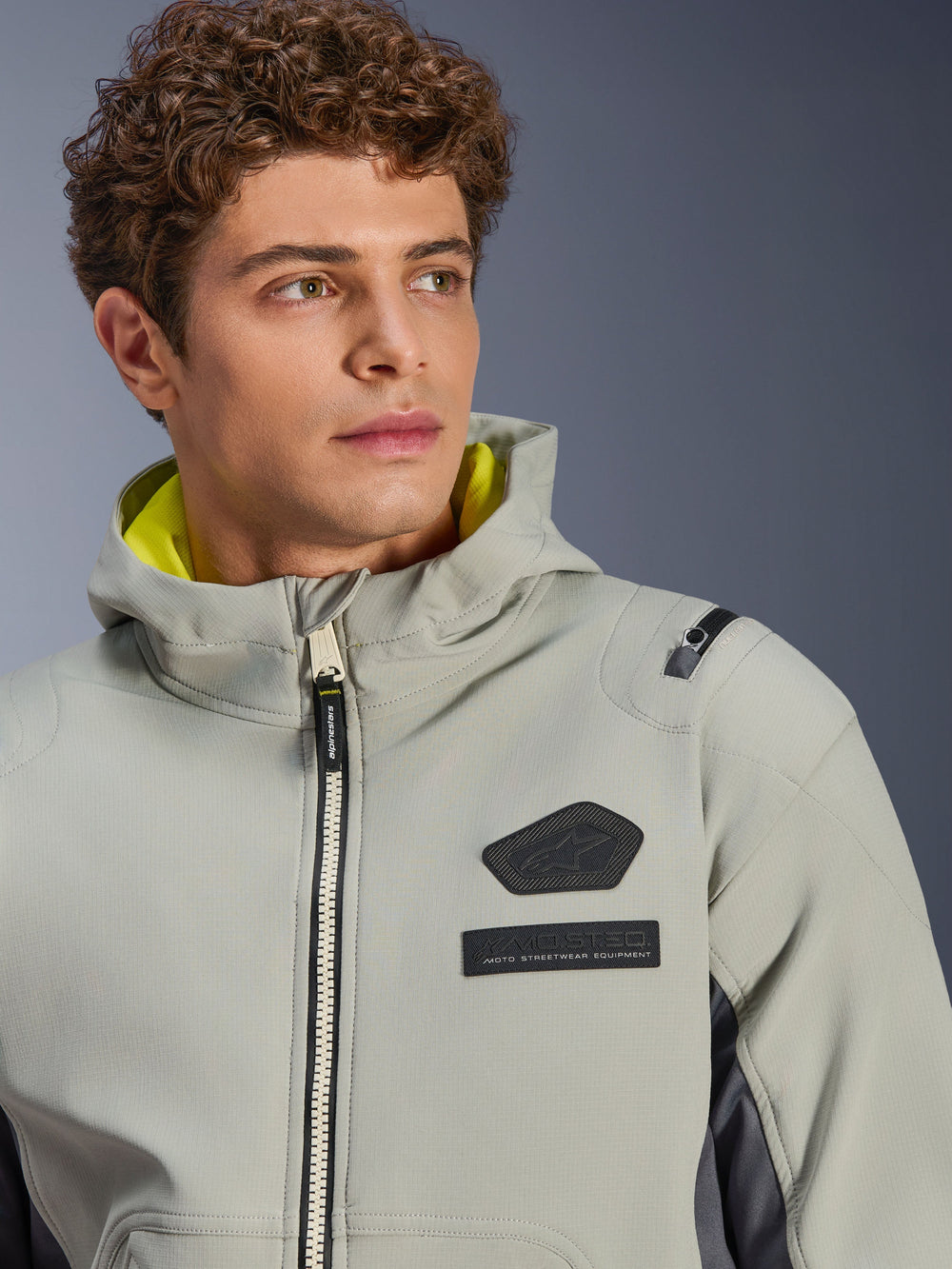 Alpinestars Moblast Waterproof Jacket, Iron Gray Dark Sage, close-up shot of male model wearing the jacket, featuring black Alpinestars logo and MO.ST.EQ. chest patches, bright yellow hood lining, and a modern technical design.
