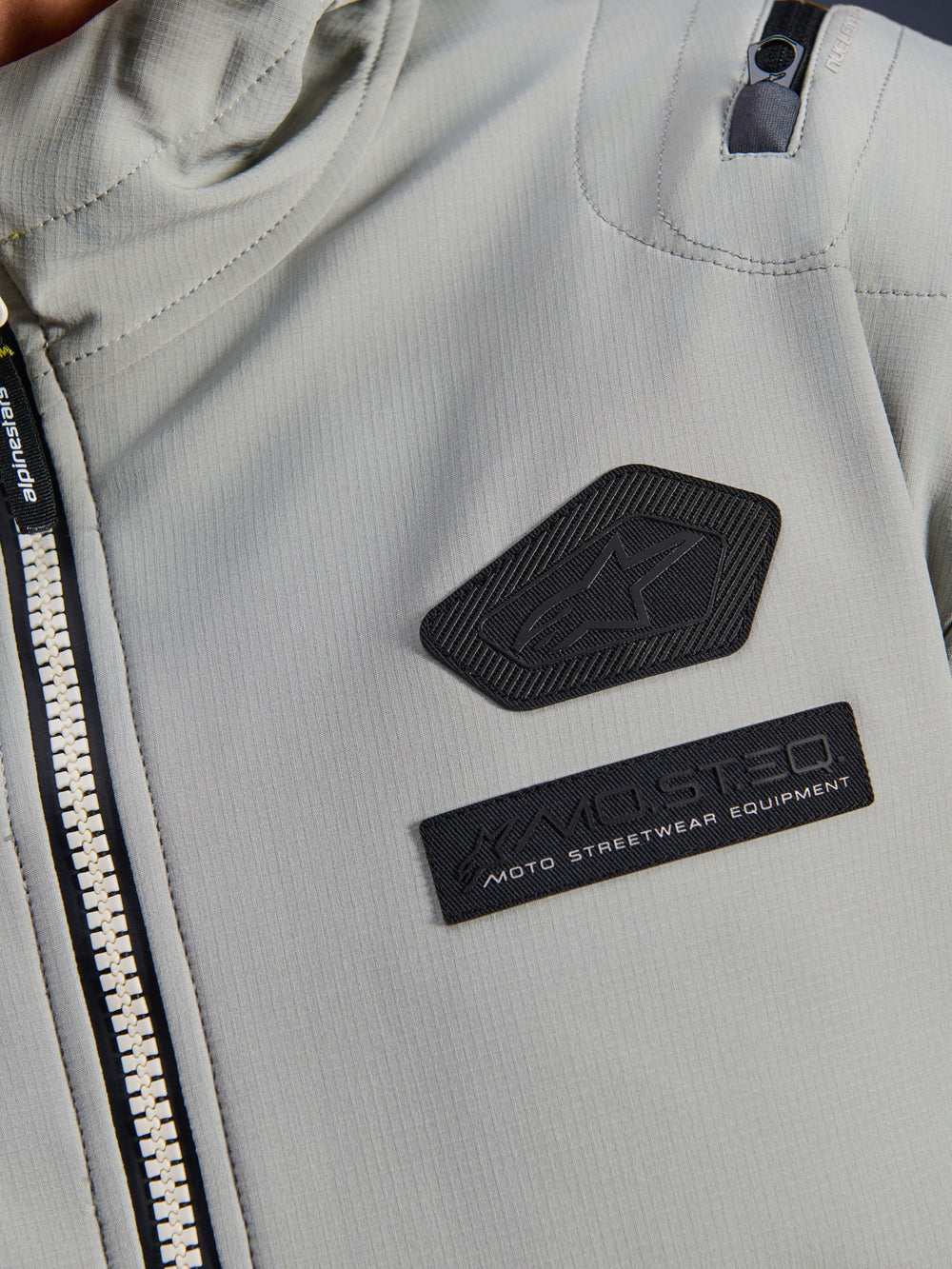 Alpinestars Moblast Waterproof Jacket, Iron Gray, close-up detail of chest patches featuring the Alpinestars logo and MO.ST.EQ. Moto Streetwear Equipment branding, technical ripstop fabric with heavy-duty front zipper and shoulder pocket detail.