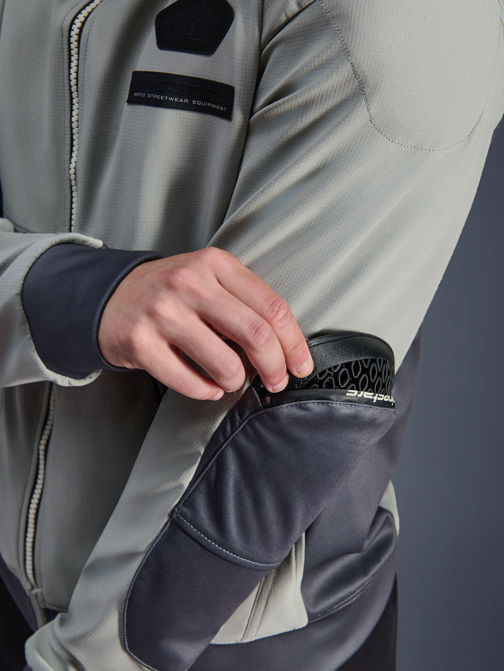 Alpinestars Moblast Waterproof Jacket, Iron Gray and Dark Sage, close-up showing removal of Nucleon Flex Plus armor from external elbow zipper pocket, technical textile urban motorcycle gear with MO.ST.EQ. branding
