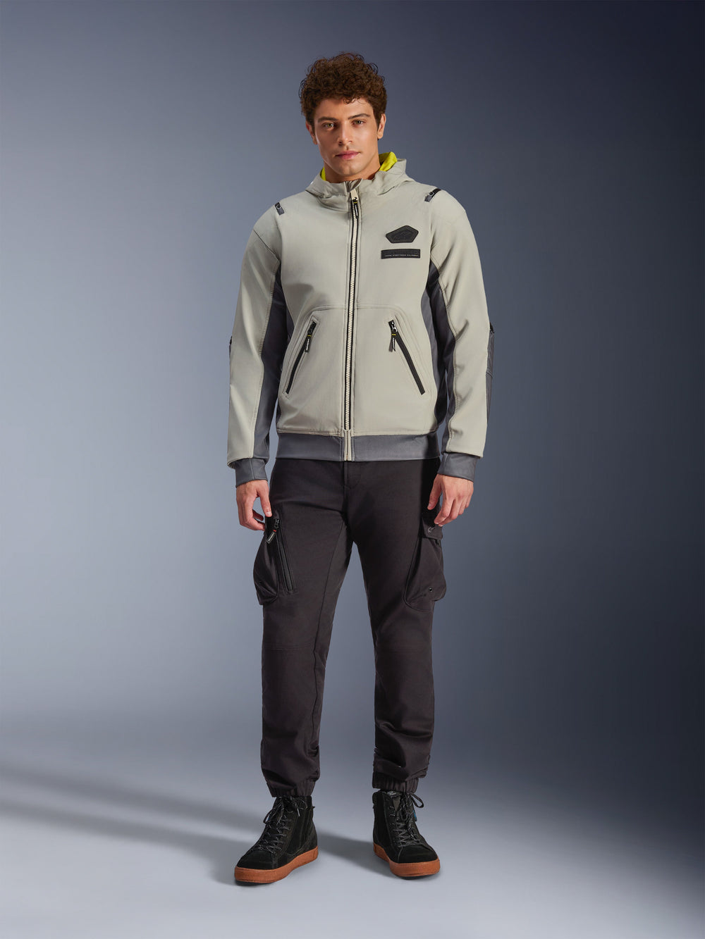 Male model wearing Alpinestars Moblast Waterproof Jacket in Iron Gray and Dark Sage, hooded motorcycle jacket with a modern technical design, paired with dark cargo riding pants and black boots, urban contemporary motorsport apparel
