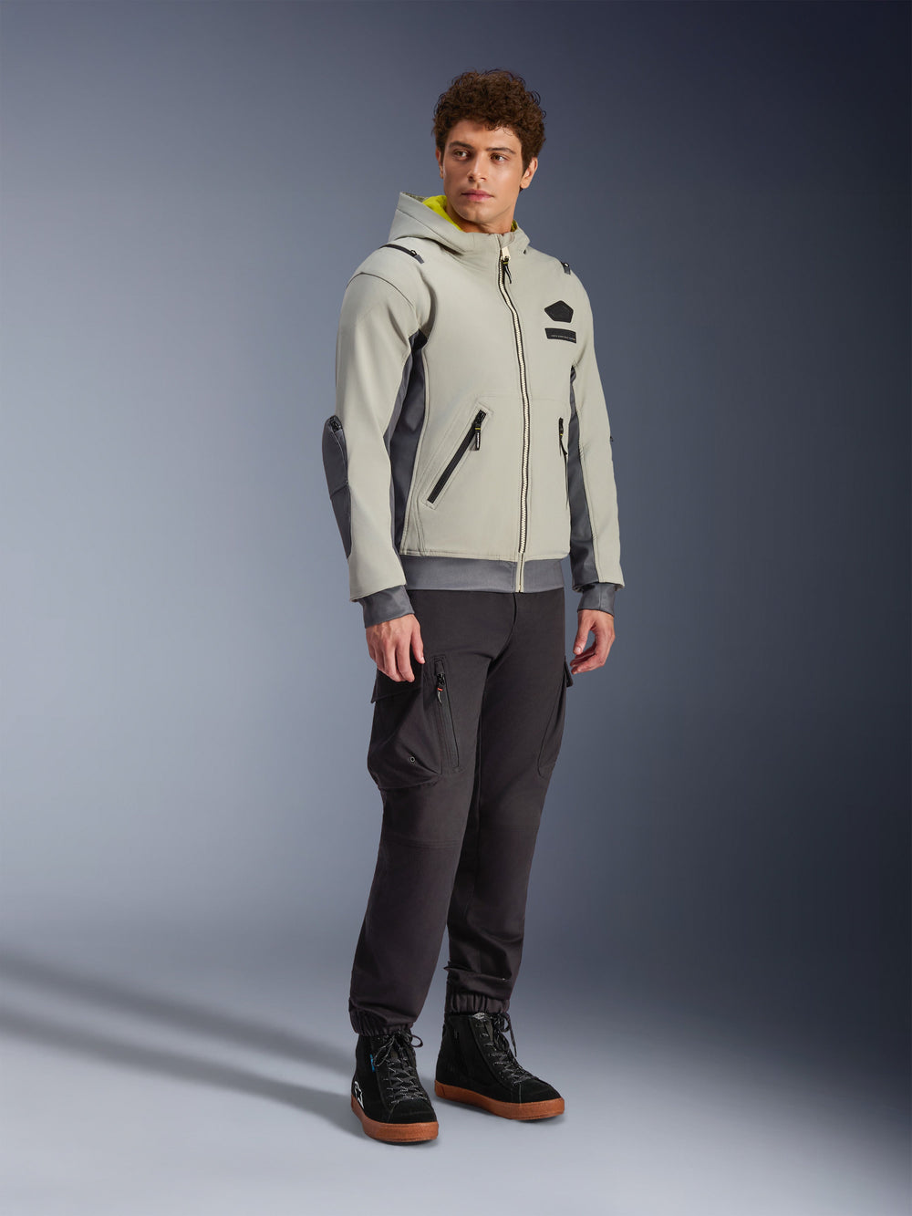 Alpinestars Moblast Waterproof Jacket, Iron Gray and Dark Sage, urban motorcycle riding jacket with a hoodie design, worn by a model with dark riding pants and black boots, minimalist cyber-tech aesthetic with a yellow interior hood and waterproof zippers