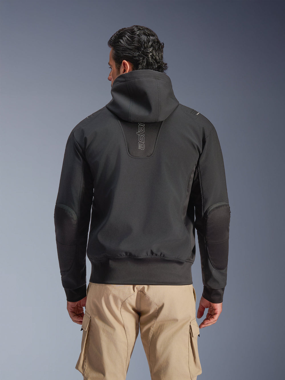 Alpinestars Moblast Waterproof Jacket, Black, Rear view of urban hooded motorcycle jacket, featuring vertical 'astars' logo on the back, elasticated waist and cuffs, reinforced elbows, and modern tech-forward design, worn with tan cargo pants.