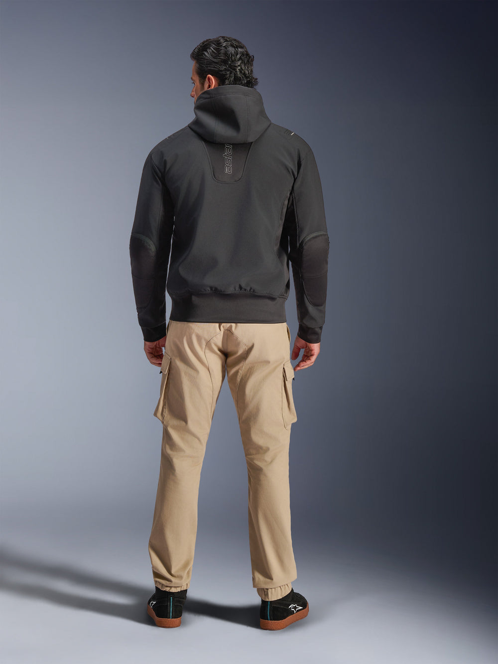 Alpinestars Moblast Waterproof Jacket, Black, back view of urban motorcycle hoodie, worn with tan cargo pants and black sneakers, minimalist design with vertical 'astars' branding on the upper back, contemporary all-weather riding gear