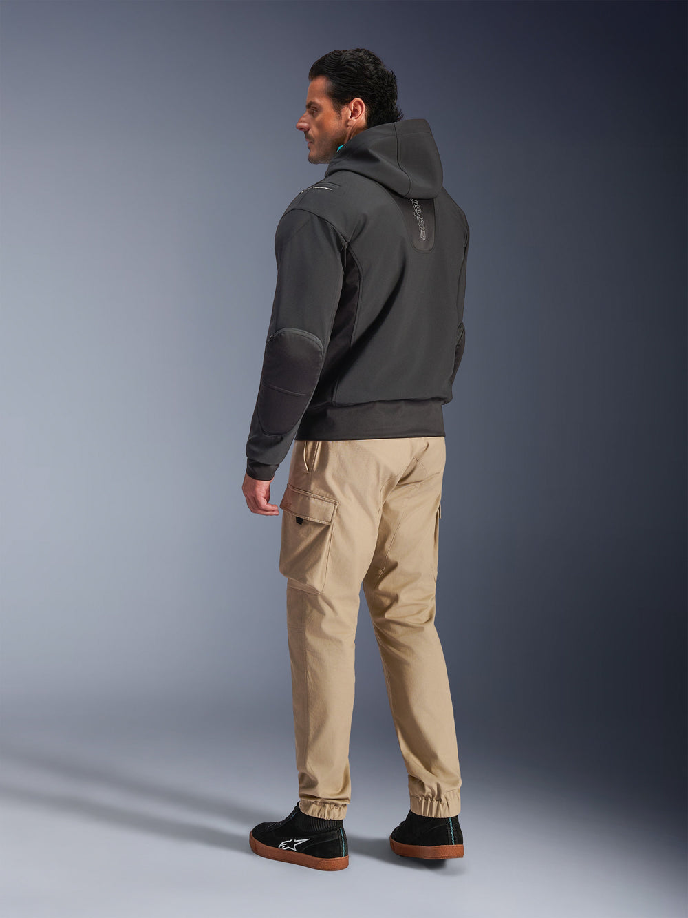 Alpinestars Moblast Waterproof Jacket, Black motorcycle tech hoodie, side and back view, worn with tan cargo pants and black sneakers, featuring a hooded design, technical textile construction, and a modern urban aesthetic.