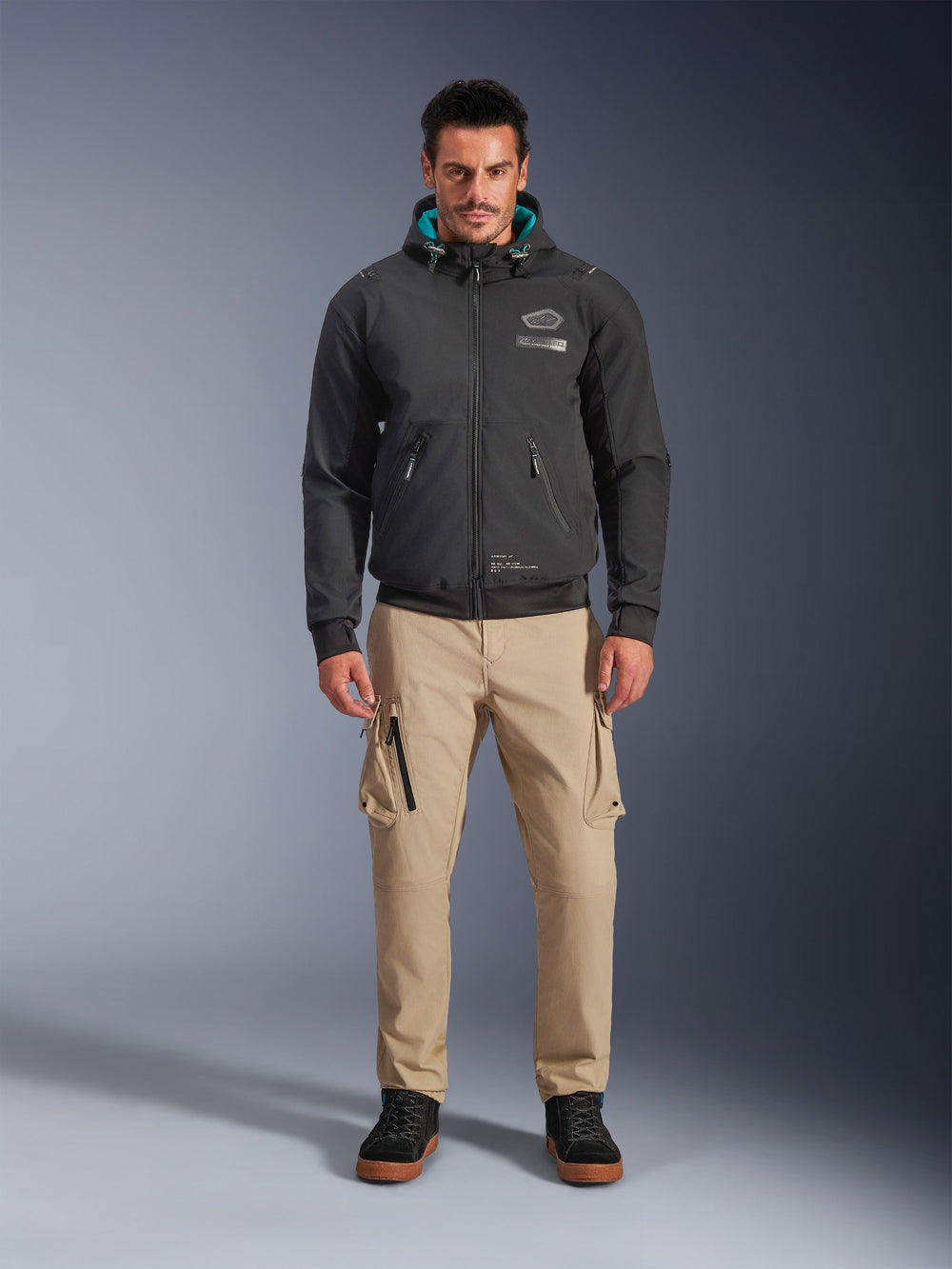 Alpinestars Moblast Waterproof Jacket, Black urban motorcycle hoodie with teal interior hood lining, worn with tan cargo pants and black high-top boots, contemporary all-season riding apparel
