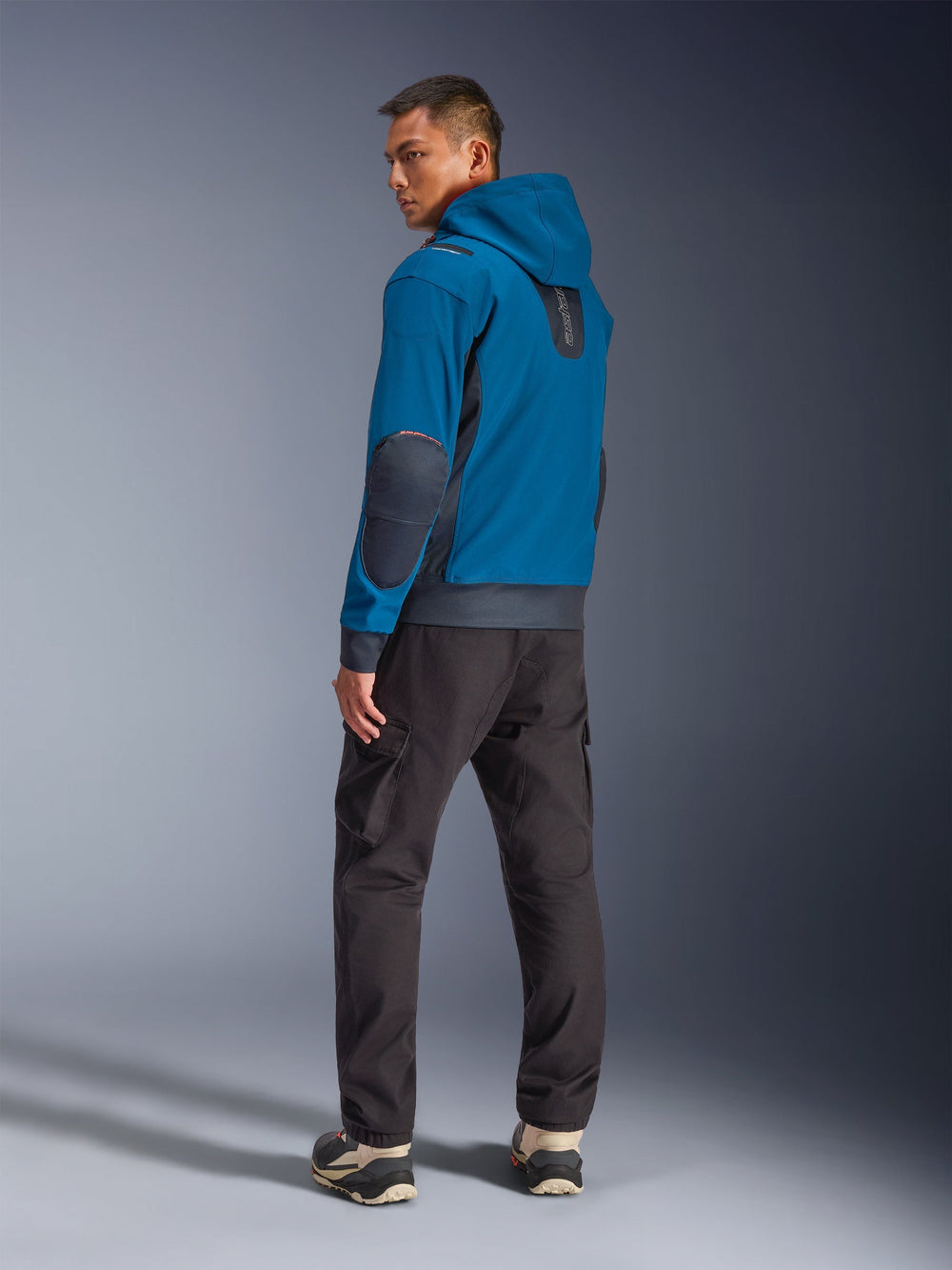Alpinestars Moblast Waterproof Jacket in Dark Petrol and Dark Navy, back view on model showing hooded design, dark navy elbow patches and side inserts, paired with dark cargo pants and technical sneakers, modern urban motorcycle apparel.
