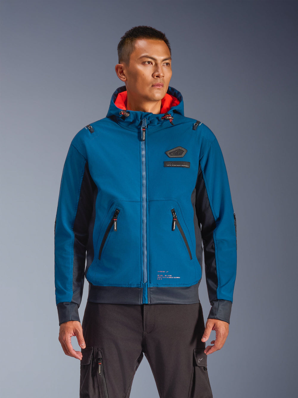 Alpinestars Moblast Waterproof Jacket worn by model, Blue and Dark Navy, Motorcycle Urban Riding Jacket, Hooded design with red interior lining, Black zippers and chest patch detail, Streetwear-inspired motorcycle gear