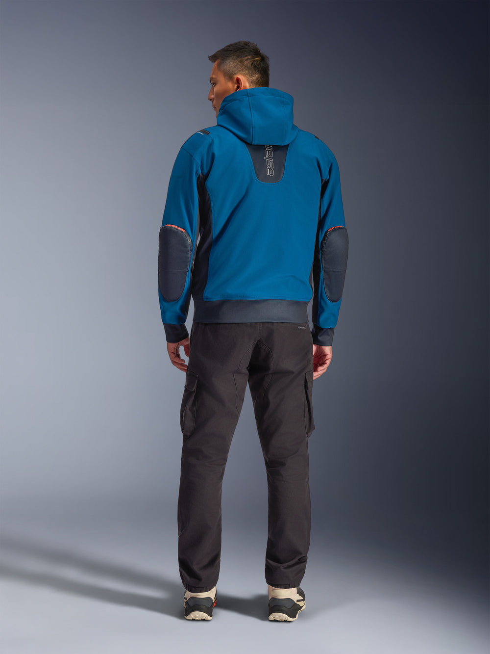 Alpinestars Moblast Waterproof Jacket, Urban Motorcycle Jacket, Dark Petrol and Dark Navy Blue, rear view showing hood and reinforced elbow patches, worn with dark cargo pants, sleek all-weather commuter riding gear