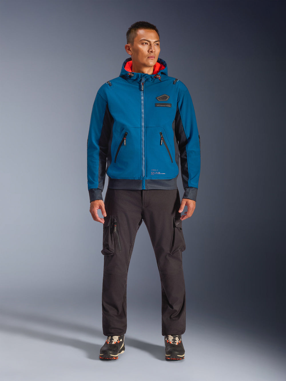 Alpinestars Moblast Waterproof Jacket, Dark Petrol and Dark Navy blue hooded motorcycle jacket with orange interior lining, worn by a male model with dark cargo pants and black sneakers, modern urban riding gear