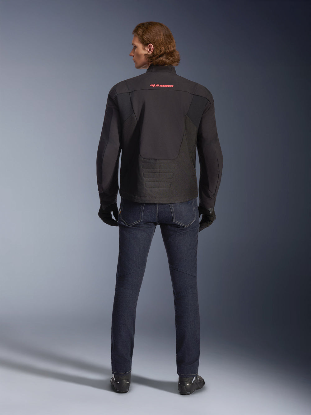 Alpinestars SMX Waterproof Jacket, Black, back view worn by a model, featuring a red logo on the upper back, elongated rear hem for riding coverage, styled with dark blue motorcycle jeans and black gloves.