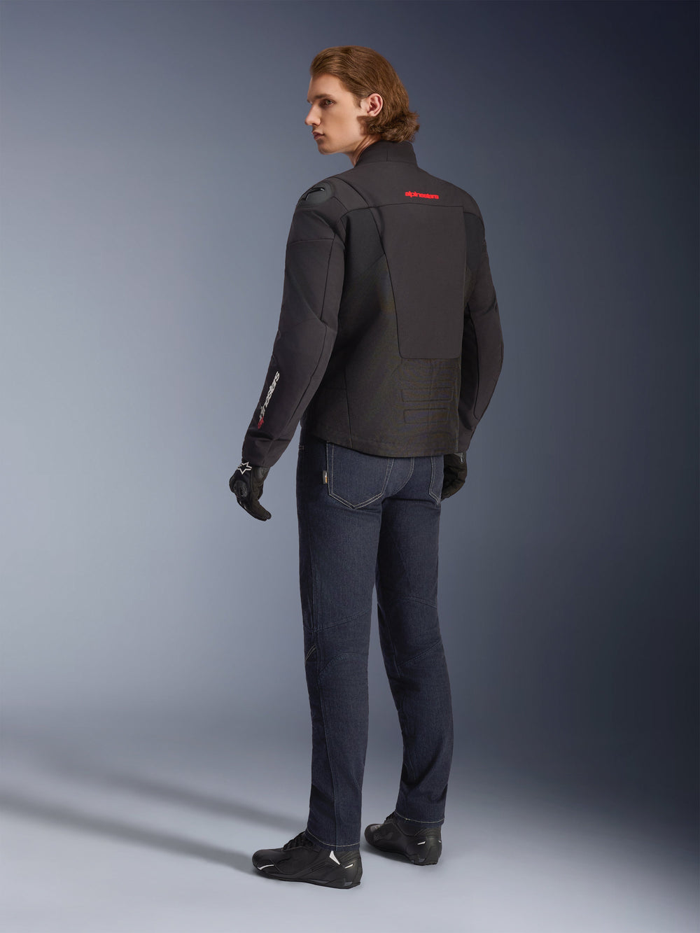 Alpinestars SMX Waterproof Jacket, Black motorcycle jacket with red and white accents, male rider shown from the back wearing dark blue riding jeans and black gloves, featuring external shoulder protection and elongated rear hem for road riding coverage