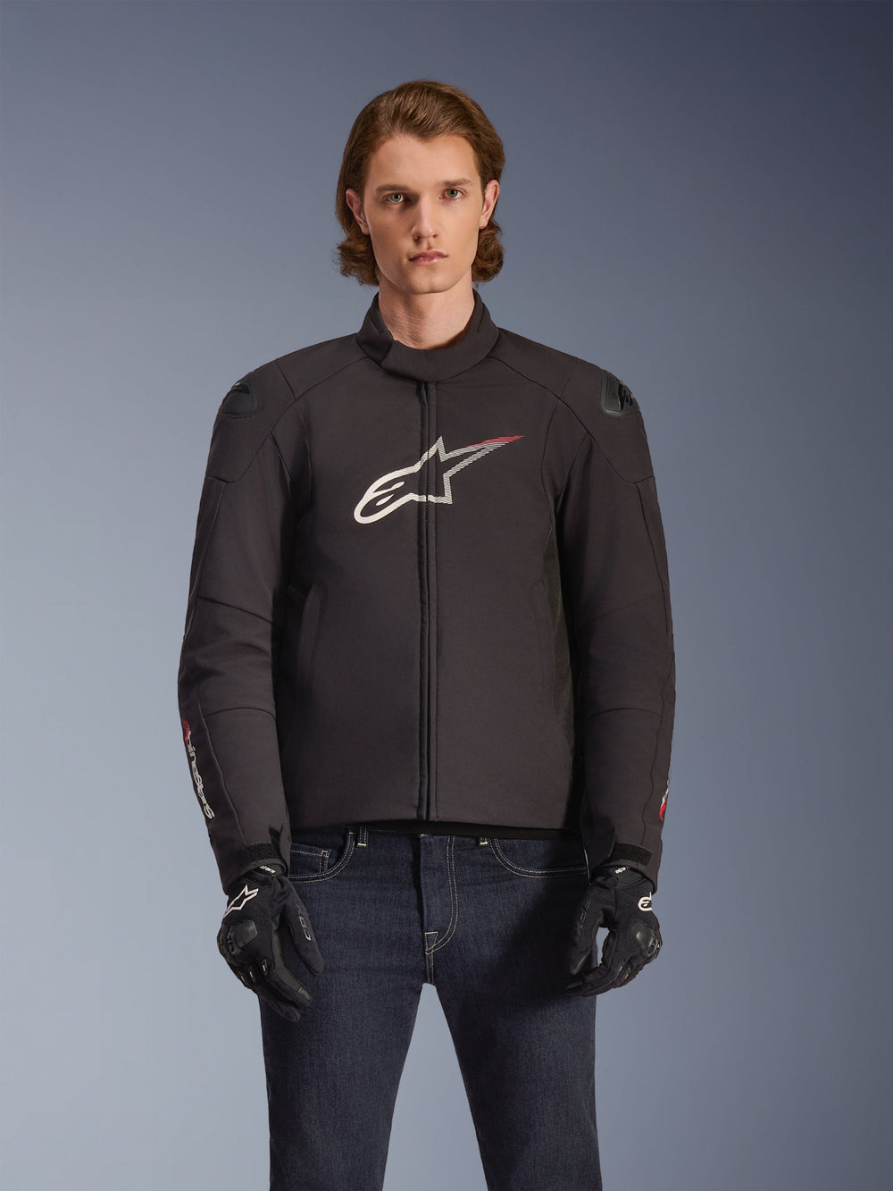 Alpinestars SMX Waterproof Jacket, Black textile motorcycle jacket with white and red star logo on the chest, worn by a model with black gloves and blue jeans, featuring external shoulder sliders and a sporty fit.