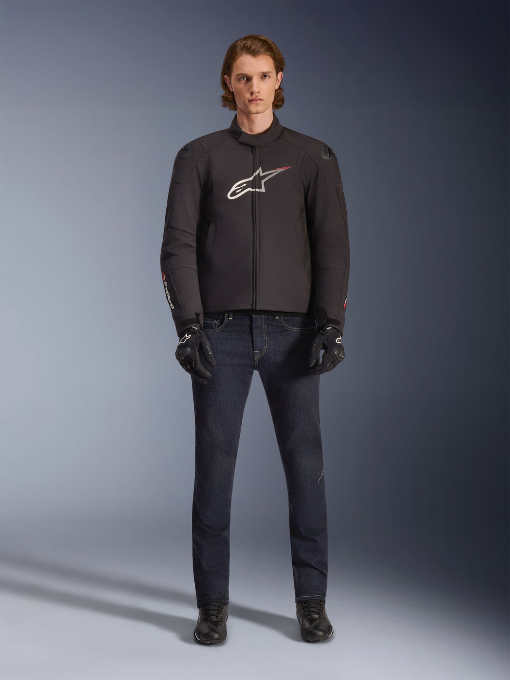 Model wearing Alpinestars SMX Waterproof Jacket in Black with white and red logo, paired with dark denim riding pants, black gloves, and riding boots, sporty all-weather motorcycle gear outfit.