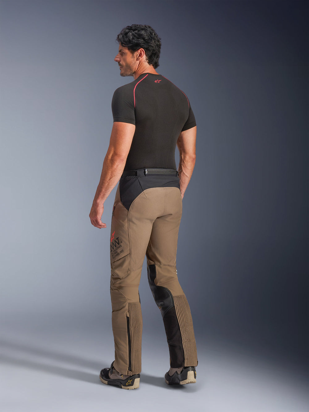 Alpinestars AMT-8 Stretch Drystar® XF Pants in Walnut Black, adventure-touring motorcycle pants shown from a back three-quarter view, featuring walnut tan textile with black panels on the seat and inner legs, accordion stretch calf panels, and AMT branding on the side, worn with a black base layer.