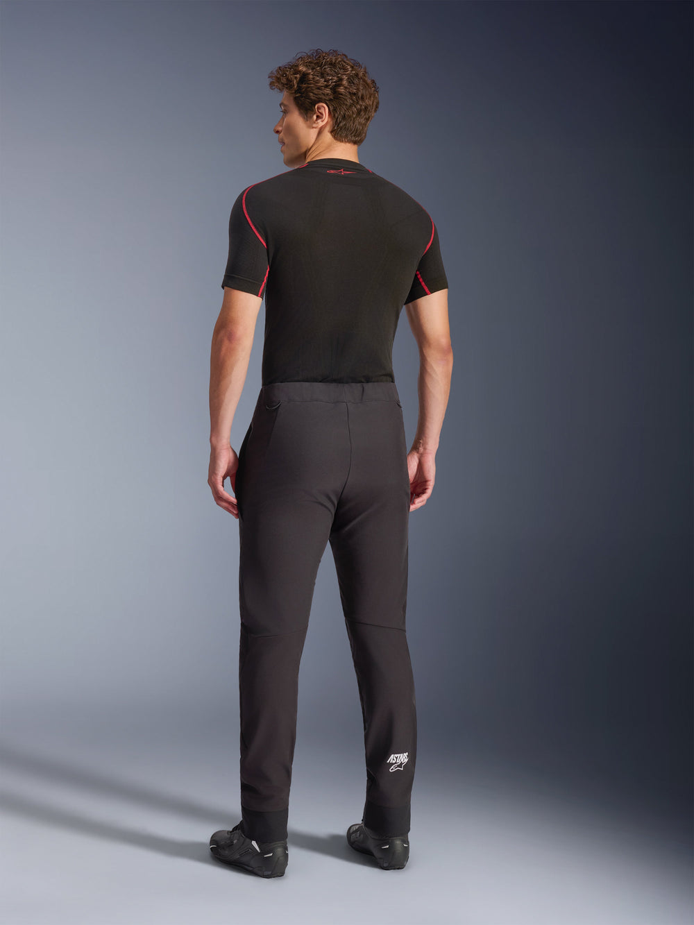 Alpinestars Aeroshell Pants, Black, softshell motorcycle pants, back view on a model, featuring a modern tapered fit, white Astars logo on the lower leg, and elasticated hems, worn with a black short-sleeved base layer and riding shoes.