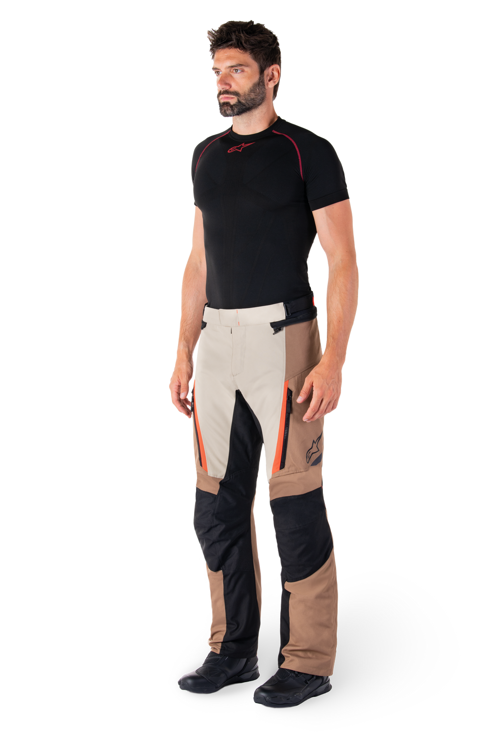 Man wearing Alpinestars ST-1 Waterproof Pants, Adventure Touring Pants, Dark Khaki Sand Black and Red Fluo, styled with a black base layer shirt and black motorcycle boots, featuring reinforced panels and orange zipper accents