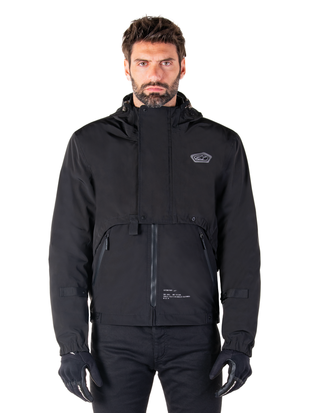 Alpinestars Mootant Waterproof Parka, Black hooded motorcycle jacket with a modular technical design, worn by a male model with black gloves, featuring a unique chest flap and zippered side pockets, urban and touring riding style.