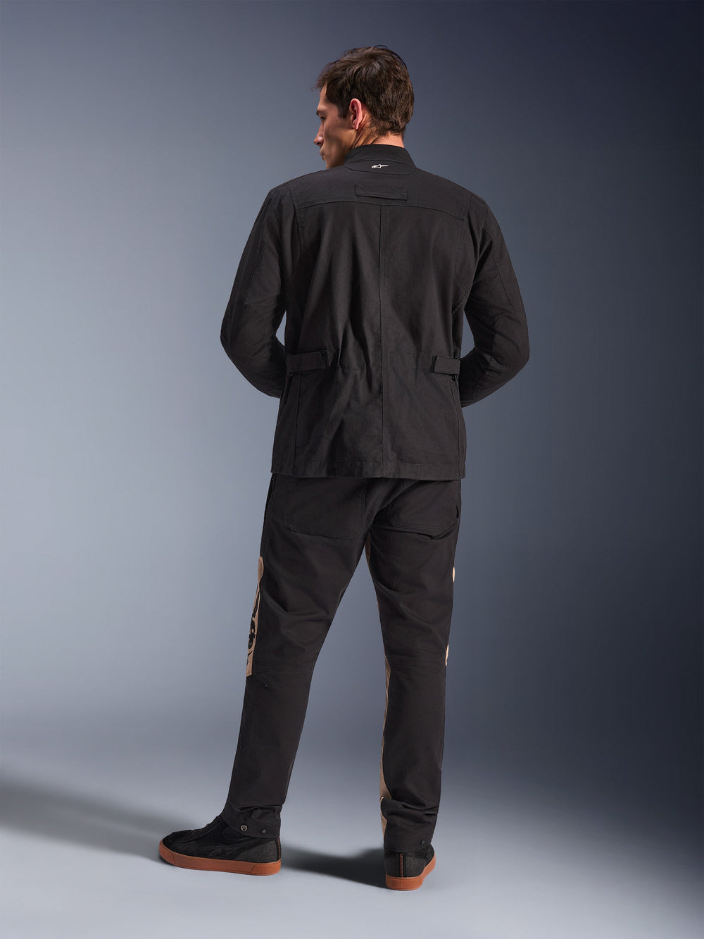 Rear view of Alpinestars Dice Jacket in Black, urban motorcycle field jacket with pleated shoulders and waist adjusters, paired with black and tan riding pants and sneakers, contemporary city commute style