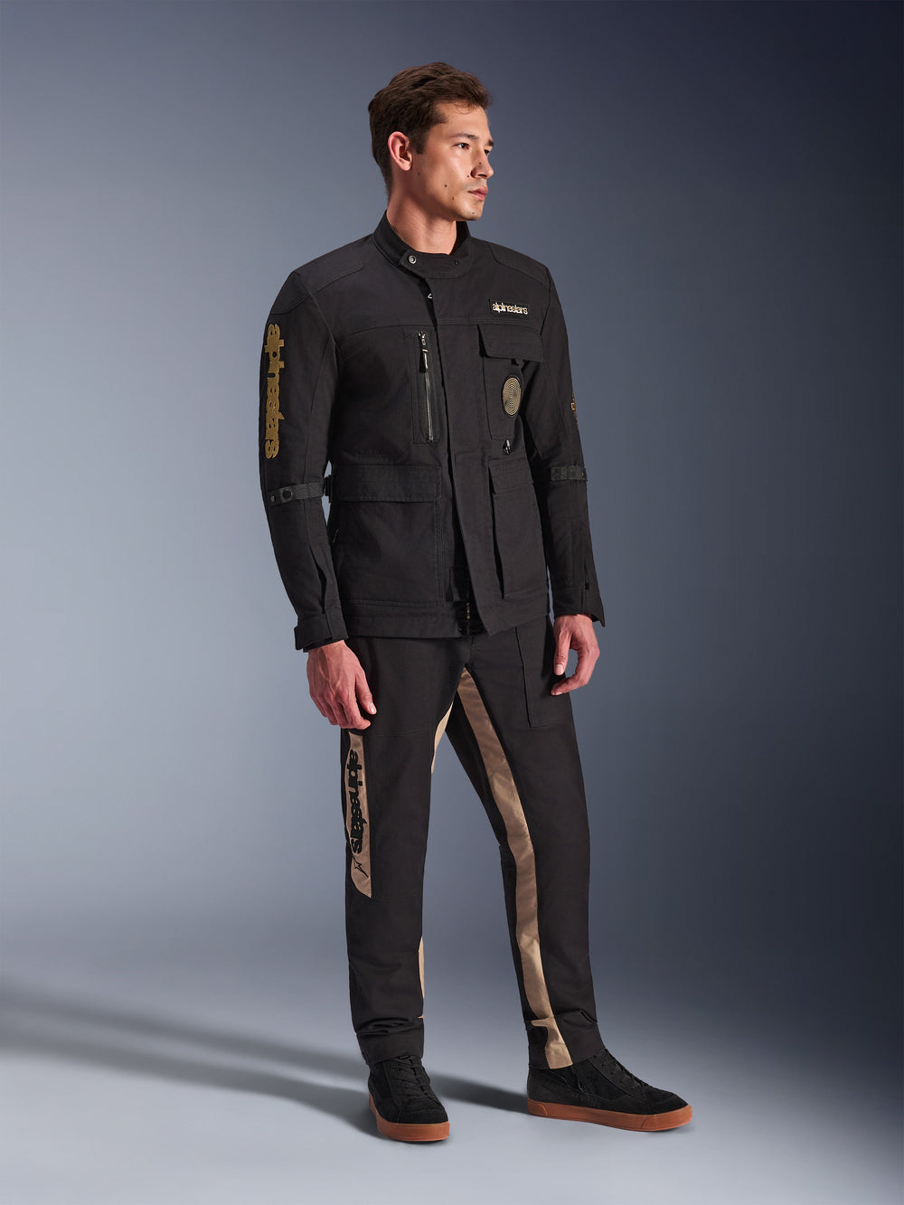 Male model wearing Alpinestars Dice Jacket in Black, urban motorcycle jacket with vintage field jacket aesthetic, featuring multiple utility pockets and gold branding on the sleeve, paired with matching black and tan pants and black sneakers.
