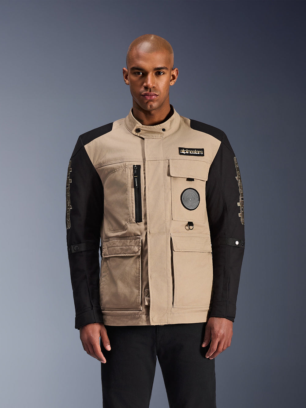 Alpinestars Dice Jacket, Urban Motorcycle Jacket, Beige and Black, rugged workwear aesthetic featuring multiple utility pockets and chest patches, high collar with button closure, bold branding on sleeves, worn by a male model