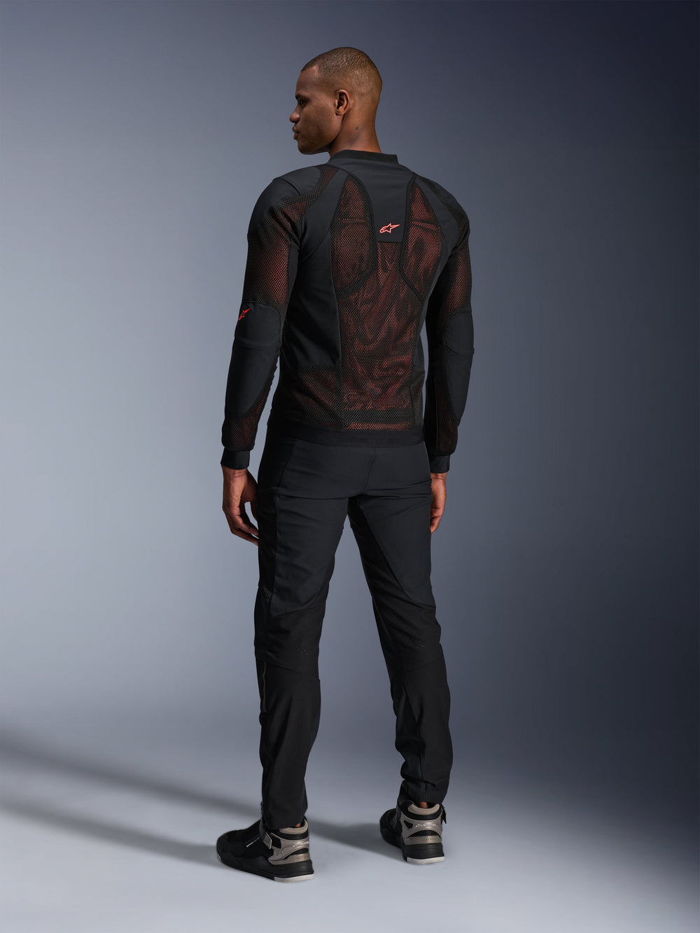 Alpinestars Troop-Air Pro Jacket in Black Red Fluo, rear view on a model, featuring a large 2D tech mesh back panel with visible red interior, paired with black technical riding pants and black/grey boots, modern urban armored motorcycle jacket design.