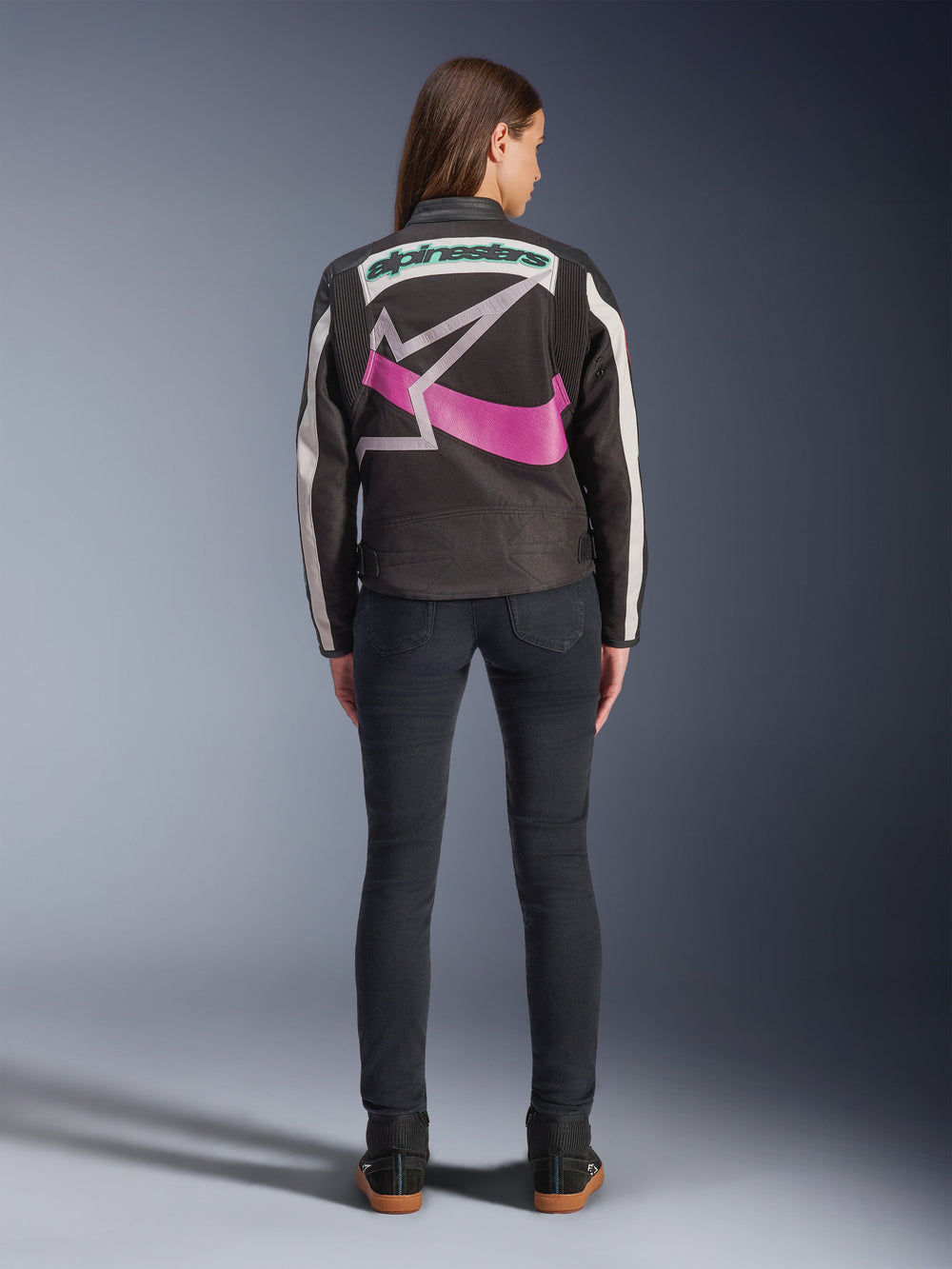 Rear view of a woman wearing the Alpinestars Idles Jacket, Black with off-white stripes, featuring a large purple and white Alpinestars star logo and green wordmark on the back, paired with dark slim-fit jeans, vintage-inspired urban motorcycle apparel
