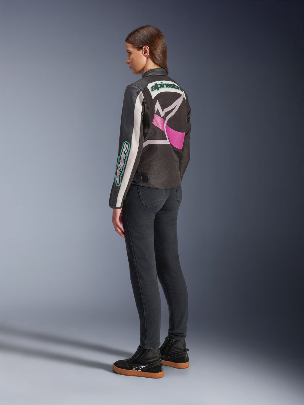 Alpinestars Idles Women’s Jacket, Motorcycle Jacket, Black Off White and Purple, viewed from the back on model, featuring a large wrap-around Alpinestars logo and vintage racing-inspired design, paired with dark jeans and black shoes.