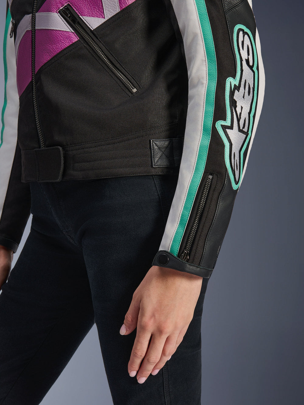 Alpinestars Idles Women's Jacket, close-up view of the sleeve and side torso, black with purple, white, and teal accents, featuring a bold Alpinestars logo on the arm, zippered cuff with snap, and waist adjustment strap, vintage-inspired motorcycle gear.