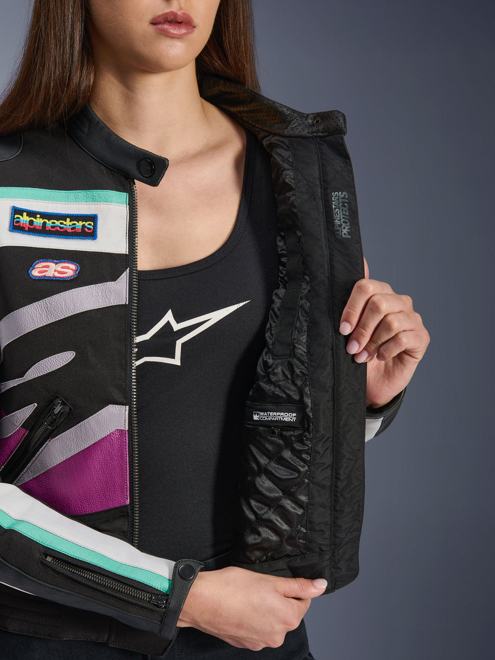Alpinestars Idles Women's Jacket, Black Off White Purple, close-up view showing the internal quilted lining with a waterproof compartment, featuring vintage-inspired racing patches and colorful teal and purple accents on the textile exterior.