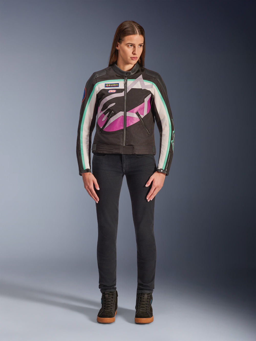 Woman wearing Alpinestars Idles Women’s Jacket, Black Off-White and Purple, vintage racing-inspired design with large wrap-around logo and teal accents, styled with black jeans and riding sneakers, urban motorcycle apparel