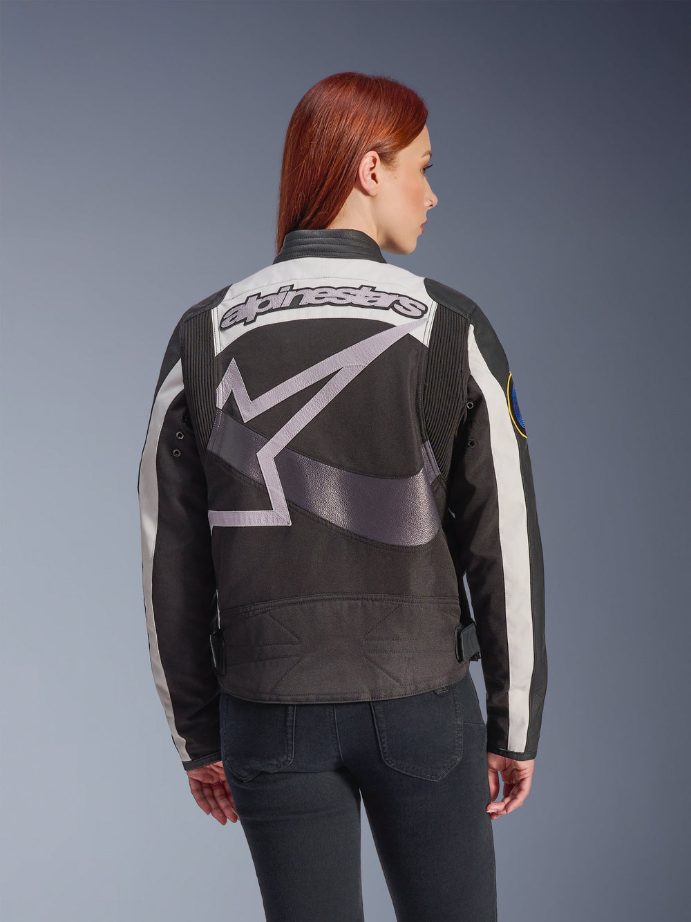 Alpinestars Idles Women’s Jacket, Black and Off White, back view featuring a large grey star logo and Alpinestars wordmark, worn by a red-haired model, vintage-inspired urban motorcycle jacket with accordion panels.