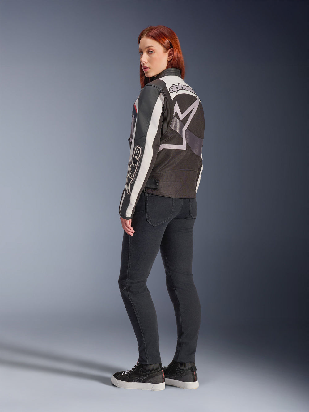 Woman wearing Alpinestars Idles Jacket, back view, Off White Flame Red and Black, featuring a large star logo on the back and Alpinestars wordmark on the sleeve, paired with black slim-fit pants and sneakers, urban motorcycle apparel for women
