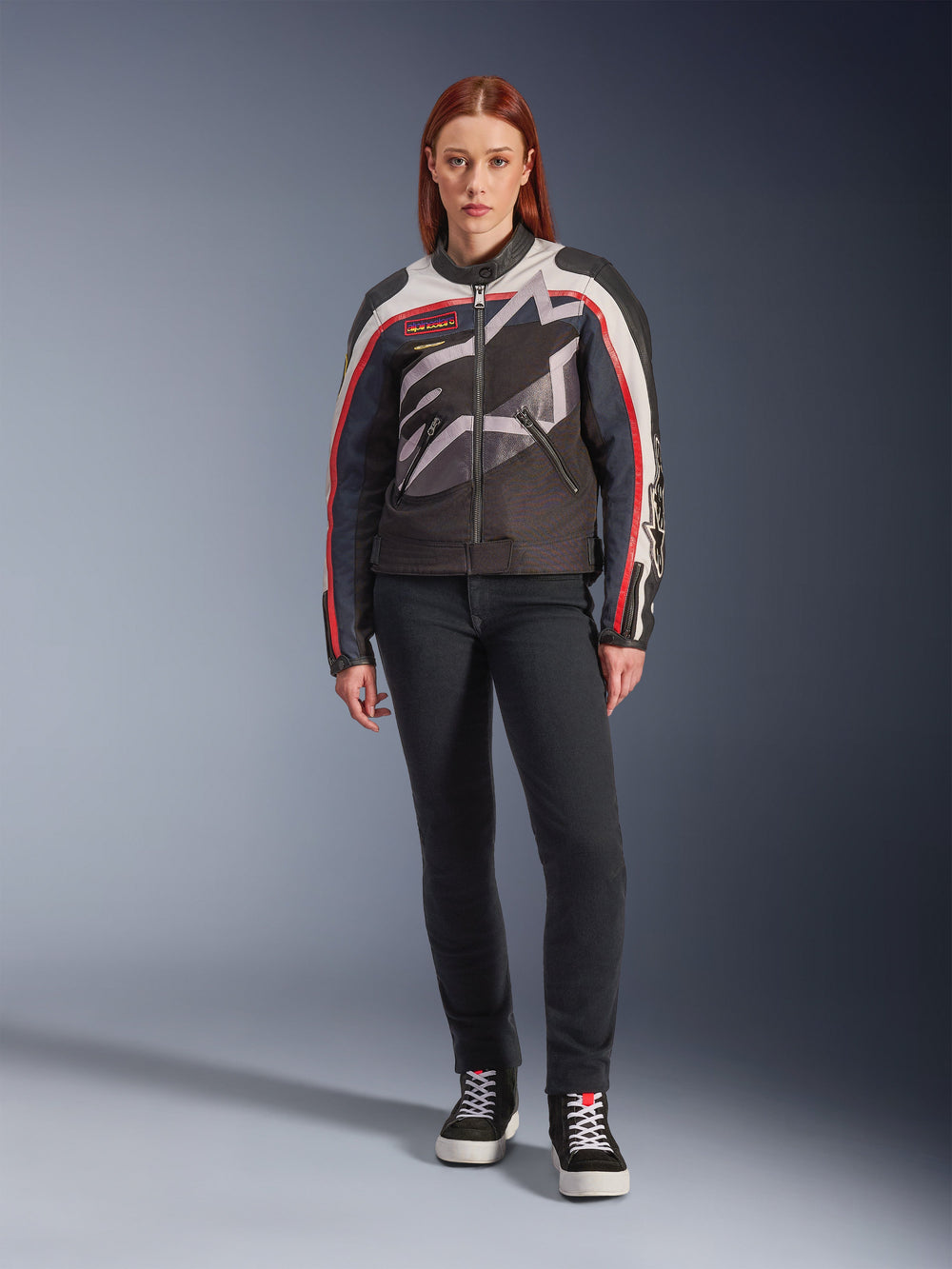 Female model wearing the Alpinestars Idles Jacket, Women's Motorcycle Jacket, Off White Flame Red and Black, featuring a large wrap-around star logo and vintage-inspired branding, paired with dark slim-fit riding pants and black high-top sneakers, urban contemporary motorcycle gear