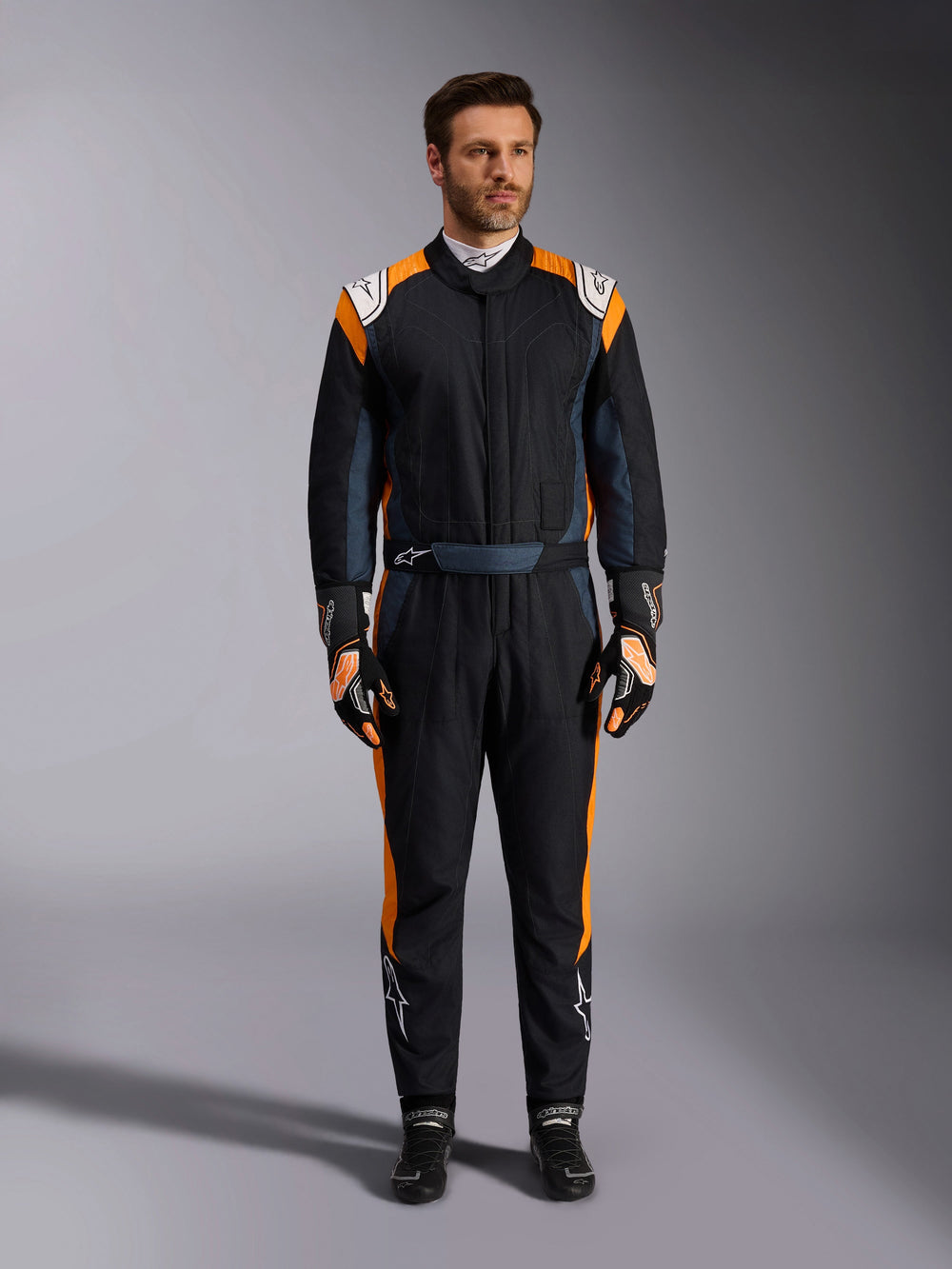 Alpinestars GP Pro Comp V2 FIA Suit, Black Asphalt and Orange Fluo auto racing suit, male model wearing full protective gear with matching racing gloves and shoes, features white Alpinestars logos and vibrant orange side panels, professional motorsport apparel