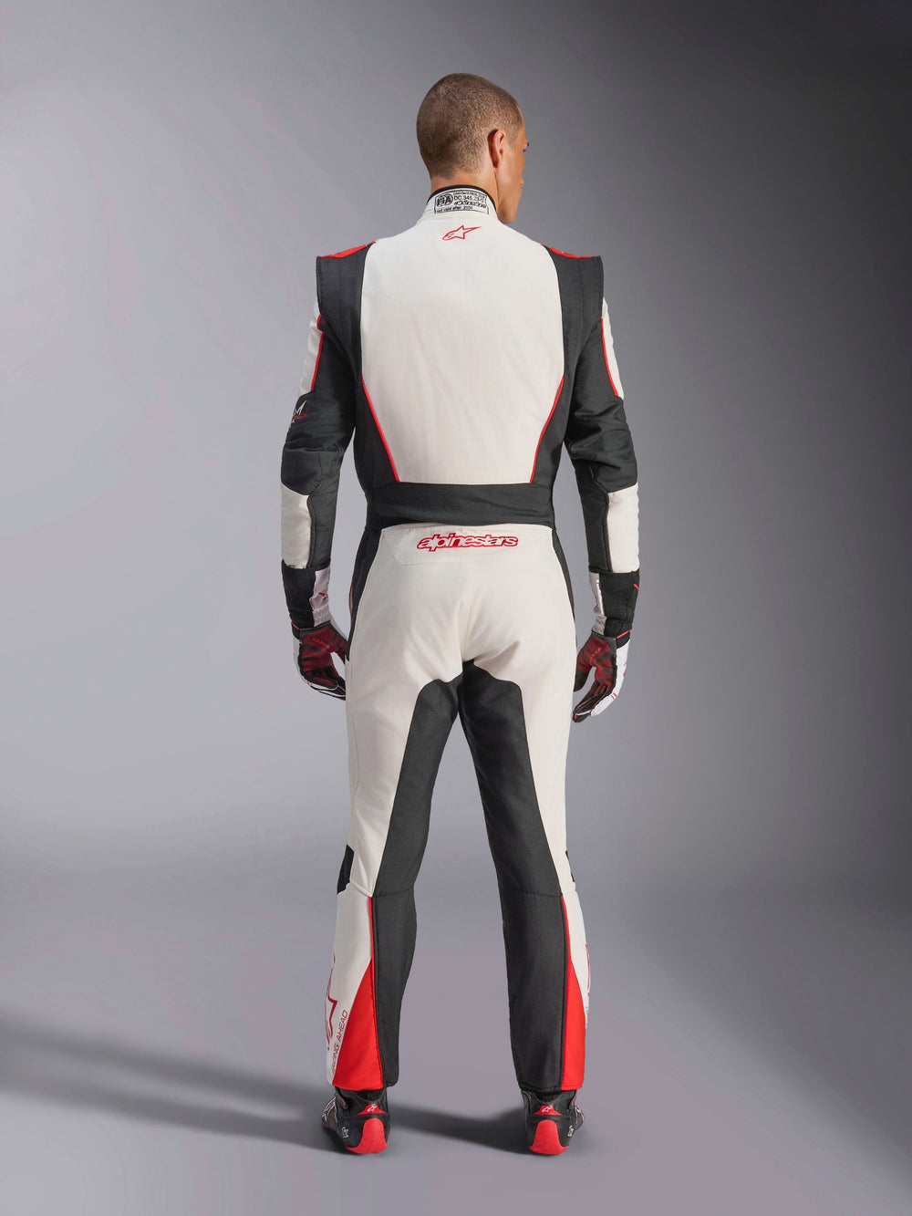 Alpinestars GP Tech V4 Suit, Auto Racing Suit, White Black and Red, back view of driver wearing the one-piece suit, featuring black stretch panels on the sides and shoulders, red accents on the lower legs, Alpinestars branding on the lower back and collar, professional FIA-certified motorsport gear
