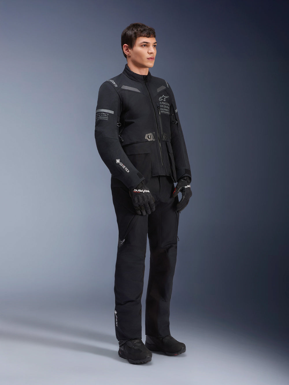 Male model wearing Alpinestars ST-7 2L GORE-TEX Jacket in Black Dark Gray, adventure touring motorcycle jacket, matching black pants and gloves, three-quarter full-body view, featuring GORE-TEX logo and Alpinestars Exploration Equipment branding