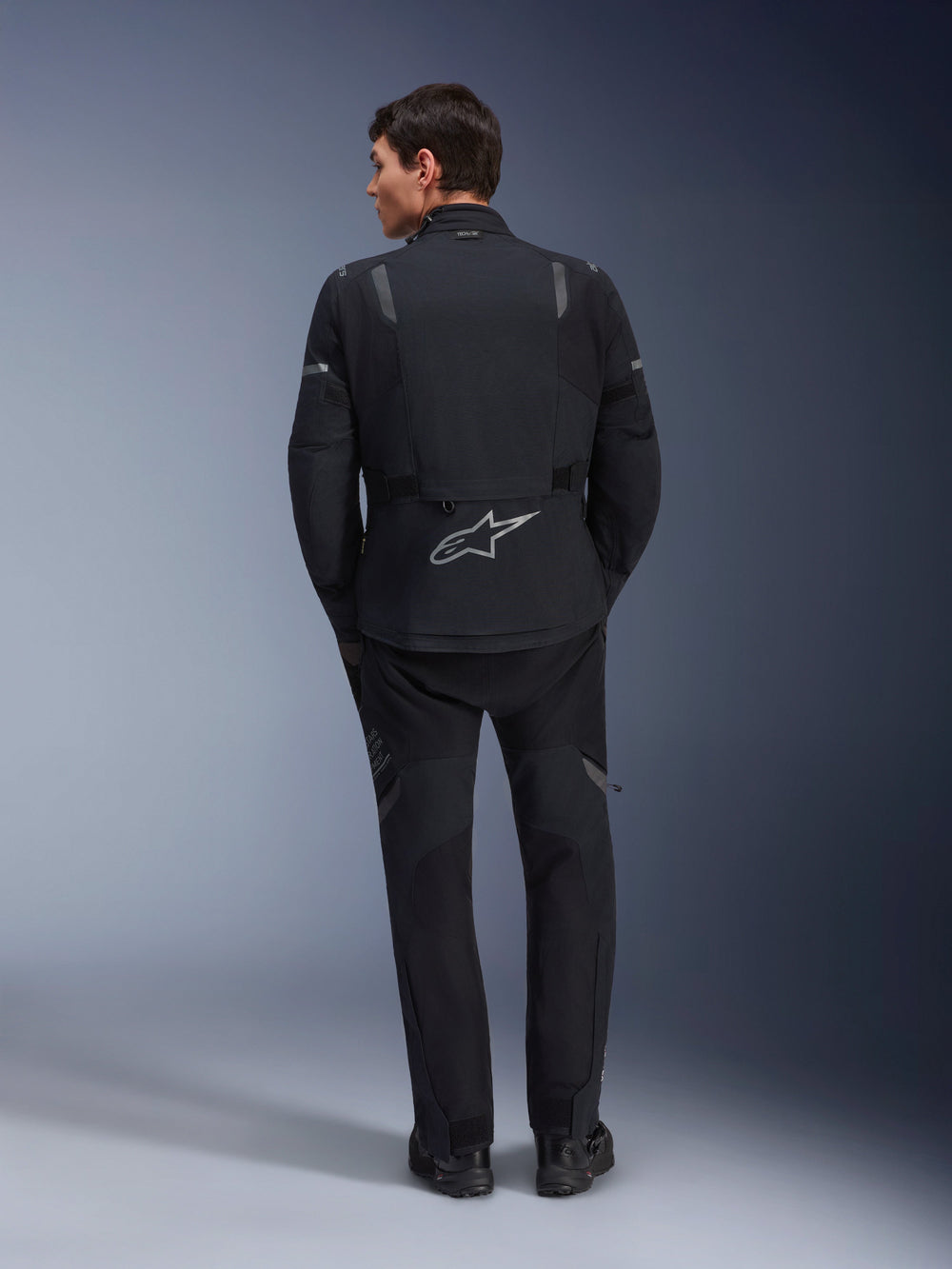 Alpinestars ST-7 2L GORE-TEX Jacket, Adventure Motorcycle Jacket, Black and Dark Gray, rear view of a man wearing the jacket and matching black pants, featuring a large reflective Astar logo on the lower back, waterproof textile gear for touring and road riding