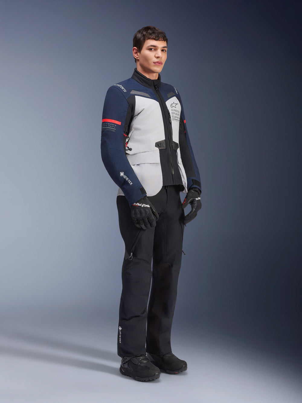 Alpinestars ST-7 2L GORE-TEX Jacket in Ice Gray, Dark Blue, and Black colorway with red accents, worn by a male model with black motorcycle pants and gloves, professional adventure touring motorcycle gear