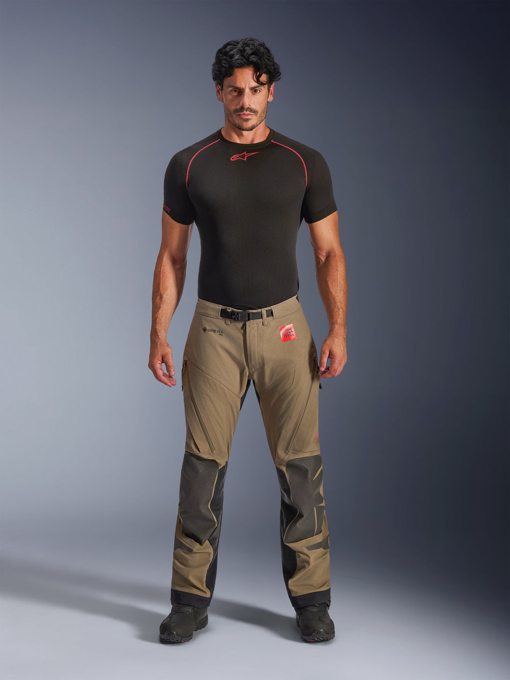 Male model wearing Alpinestars Nazca 3L GORE-TEX PRO Pants in walnut brown and black, technical adventure riding gear featuring Superfabric knee reinforcements and a magnetic waist buckle, paired with a black Alpinestars base layer shirt, studio front view.
