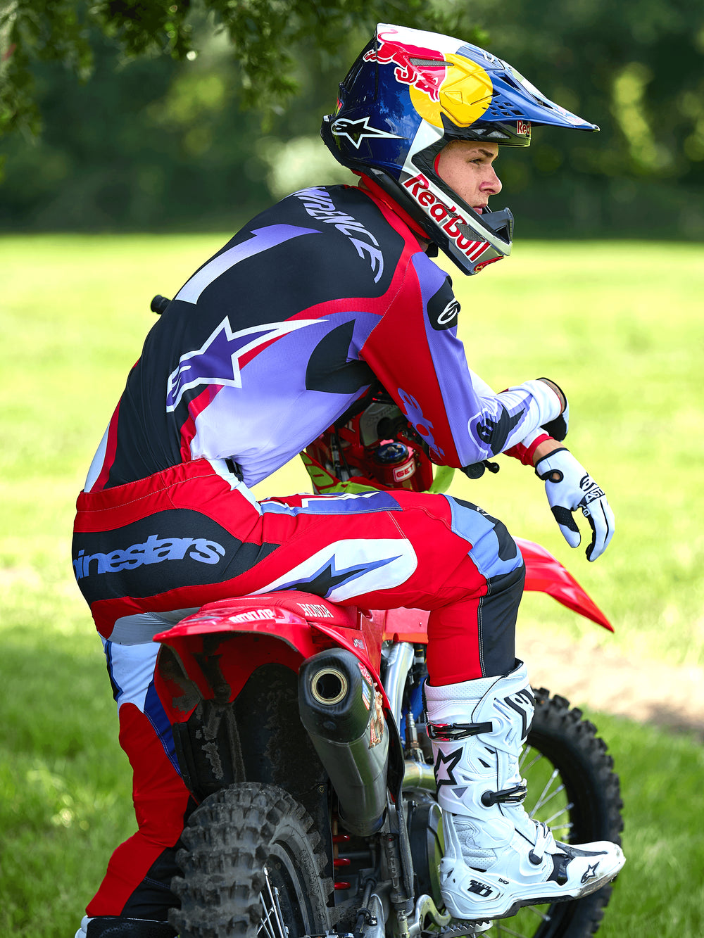 Motocross rider sitting on a red dirt bike, wearing Alpinestars 2025 Fluid Wurx Pants in Purple and Bright Red, matching jersey, white and black Tech 10 boots, white gloves, and Red Bull helmet, complete off-road racing outfit in an outdoor setting