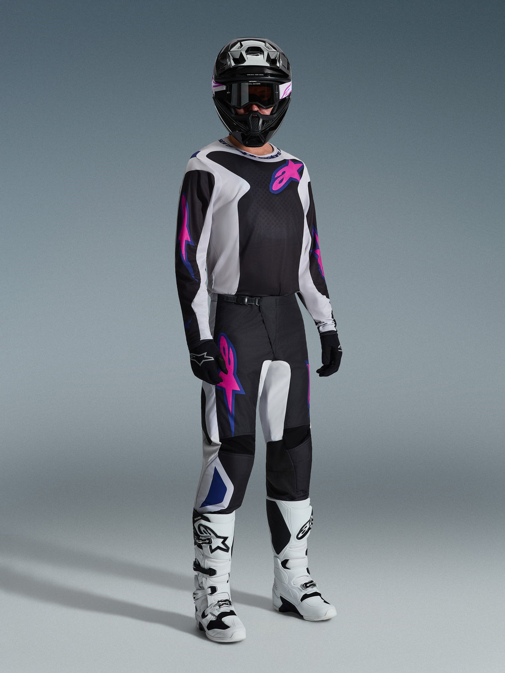 Motocross rider wearing complete racing gear, Alpinestars 2026 Fluid Grid Pants in Black Light Gray and Purple, matching jersey with pink and blue graphics, black helmet and goggles, white and black boots, and black gloves, coordinated outfit for off-road racing