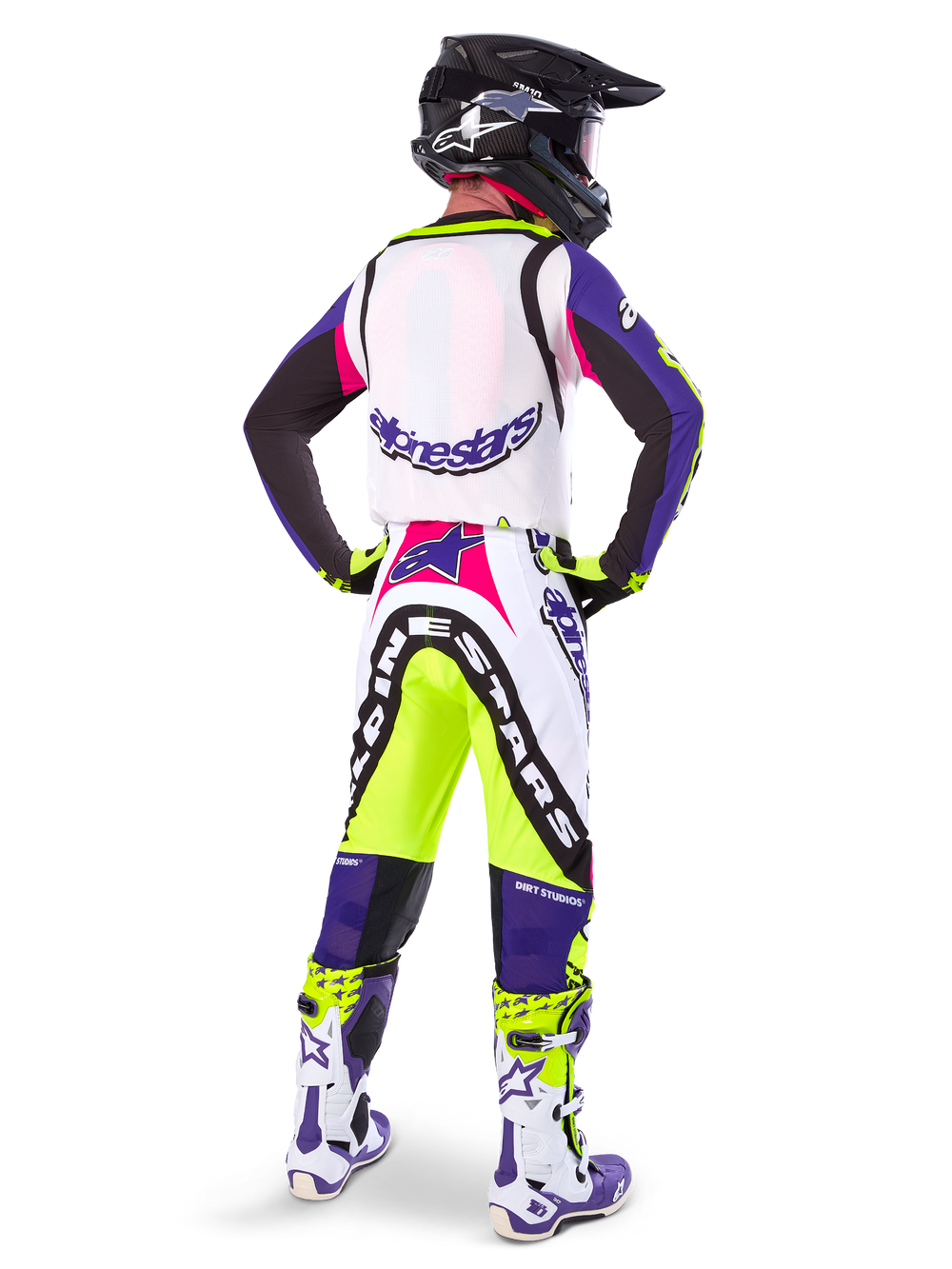 Motocross rider wearing Alpinestars Limited Edition Supertech Dirt Studios® Pants in White Purple and Yellow Fluo, seen from the back with matching jersey, boots, and black carbon helmet, professional off-road racing gear with bold graphics and neon accents.