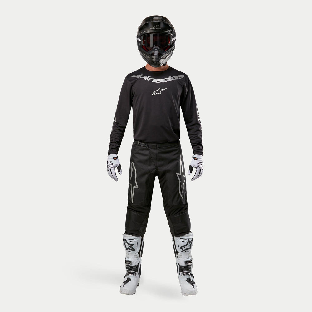Motocross rider wearing complete racing gear, Alpinestars 2024 Fluid Graphite Pants in Black and Silver, black long-sleeve jersey with silver branding, black helmet with goggles, white and black boots, white and black gloves, coordinated outfit for motocross