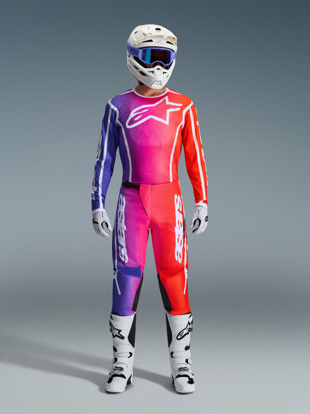 Motocross rider wearing a complete Alpinestars 2026 Fluid Apex racing gear set, featuring a purple, pink, and orange gradient jersey and pants. The coordinated outfit includes a white Alpinestars logo on the chest, a white helmet with blue goggles, and white boots, designed for off-road performance.