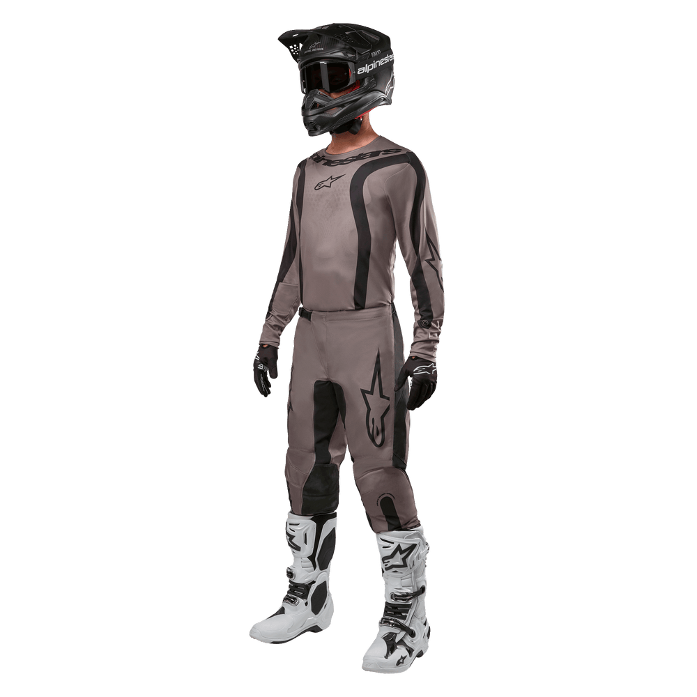 Motocross rider wearing complete racing gear featuring the Alpinestars 2024 Fluid Lurv Jersey in Mud Black, paired with matching pants, black helmet with goggles, white and black boots, and black gloves, coordinated outfit for off-road racing