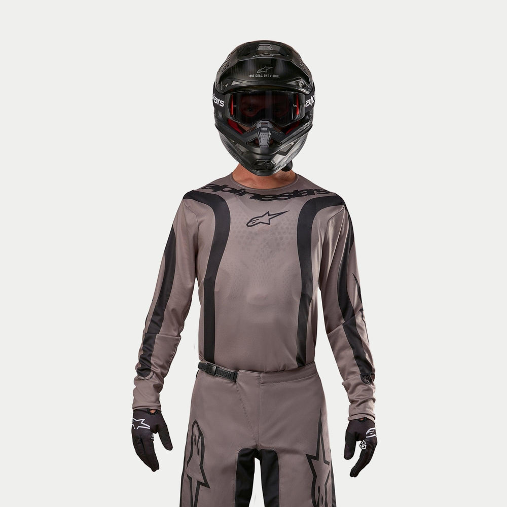 Motocross rider wearing the Alpinestars 2024 Fluid Lurv Jersey in Mud Black, an earthy grey-brown fabric with black graphic stripes and a star logo on the chest, paired with a black helmet, red goggles, and black gloves for a coordinated off-road racing look